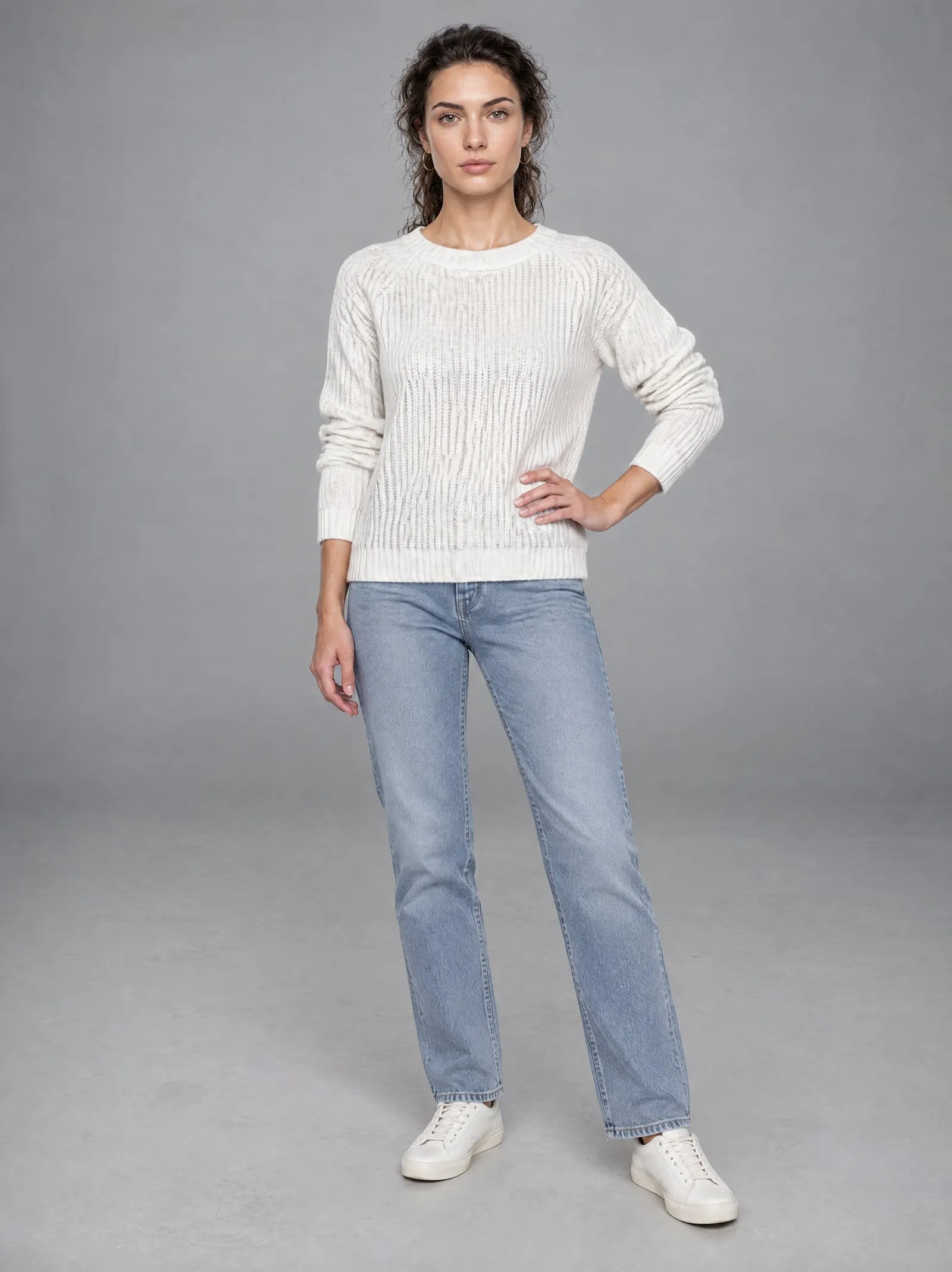 Mira wearing the white knit sweater, hand on hip, in a grey studio