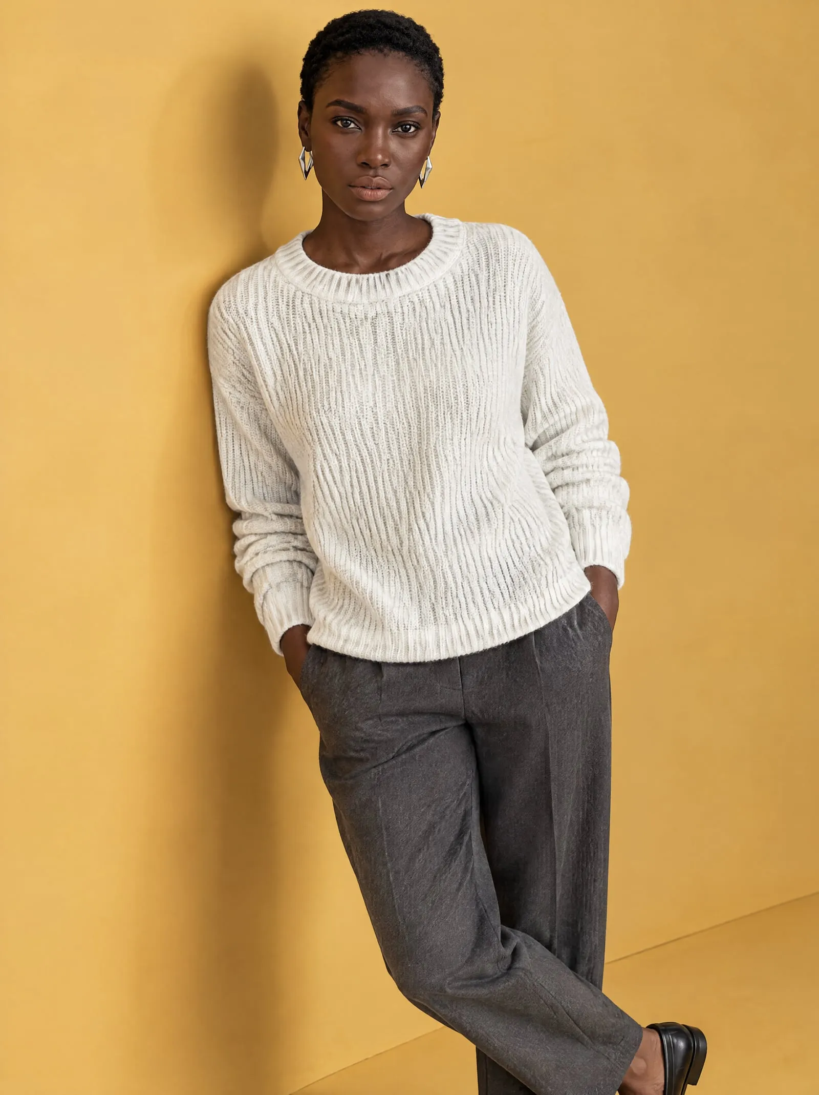 Amara wearing the white knit sweater, leaning, in a mustard-yellow studio